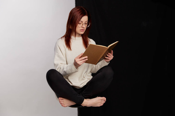 Schöne rothaarige junge Frau, die Buch liest, während sie mit überkreuzten Beinen auf einem Stuhl sitzt, isoliert über einem halb schwarz-weißen Hintergrund - Foto, Bild