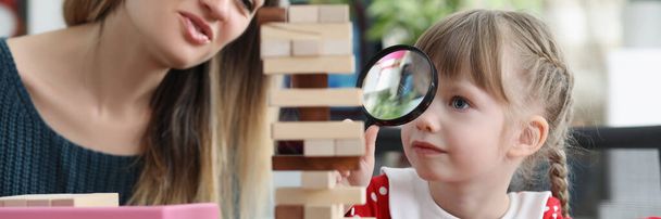 Portrait of cute little girl watch through magnifying glass on high wooden tower. Help child in development, improve concentration. Home education concept - Zdjęcie, obraz