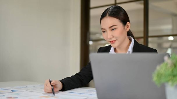 Atractiva y encantadora joven empresaria asiática o gerente ejecutiva en traje formal, trabajando en su informe de datos financieros en la oficina. - Foto, Imagen