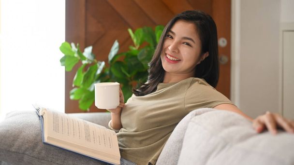 Cheerful asian woman in casual clothes holding cup of tea in her hand, relaxing on couch at home. - Foto, afbeelding