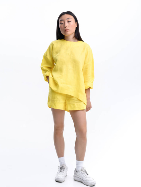 Hermosa chica asiática en un traje casual de lino amarillo posando contra una pared blanca en un estudio de fotografía - Foto, imagen