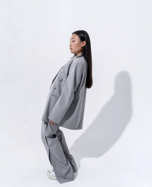 A beautiful Asian girl in a gray suit on a naked body poses against a white wall in a photo studio. Fashion shooting - Foto, immagini
