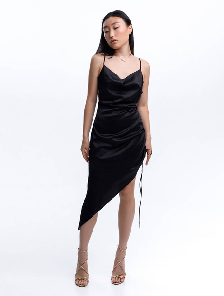 Een mooi Aziatisch meisje in een zwarte jurk poseert tegen een witte muur in een fotostudio. Mode schieten - Foto, afbeelding