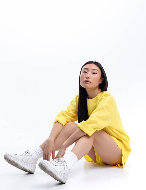 Bella ragazza asiatica in un abito casual di lino giallo in posa contro un muro bianco in uno studio fotografico - Foto, immagini