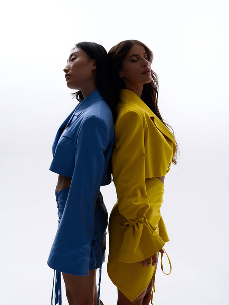 İki güzel beyaz ve Asyalı kız beyaz zeminde uzanır ve bir fotoğraf stüdyosunda mavi ve sarı takım elbiseler içinde poz verirler. - Fotoğraf, Görsel