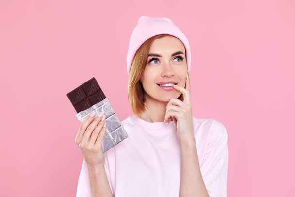 Stylish young blonde woman in t-shirt and hat holding chocolate bar on pink background - Fotoğraf, Görsel