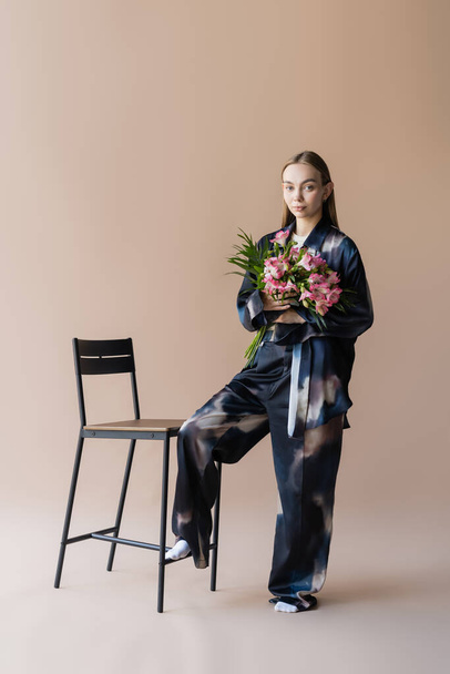 fashionable woman with alstroemeria flowers looking at camera near chair on beige background - 写真・画像