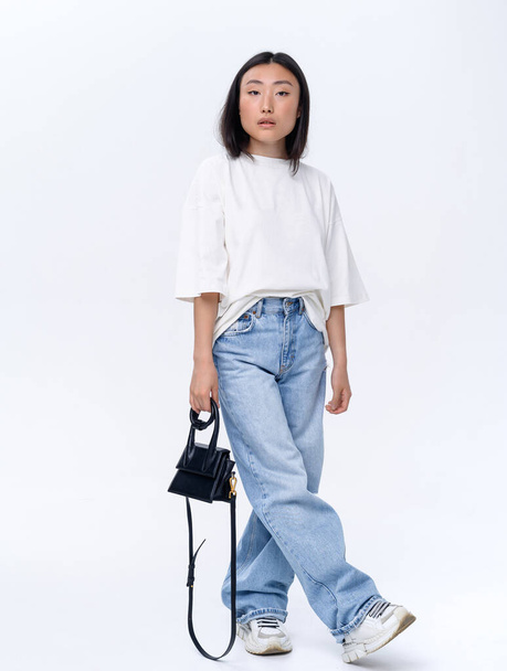 A beautiful Asian girl in a white t-shirt and blue jeans poses against a white wall in a photo studio. Fashion shooting - Foto, afbeelding