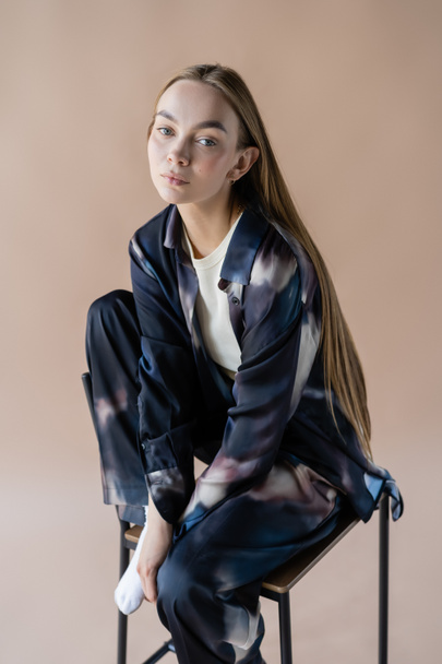 young long haired woman in tie-dye style clothes posing on chair isolated on beige - Foto, immagini
