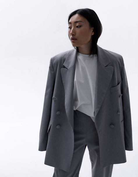 A beautiful Asian girl in a gray suit poses against a white wall in a photo studio. Fashion shooting - Photo, image