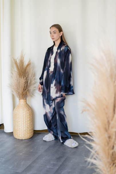 full length of woman in oversize suit and fluffy slippers near wicker vase with spikelets and white drapery - Foto, Imagen