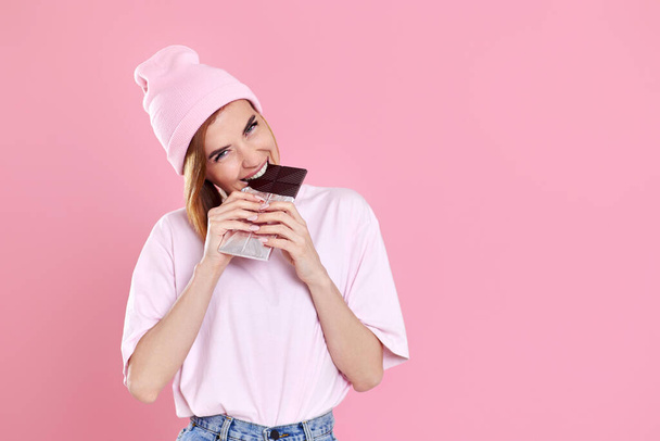 young happy woman in casual t-shirtand hat biting sweet chocolate bar isolated on pastel pink background. copy space - Fotoğraf, Görsel