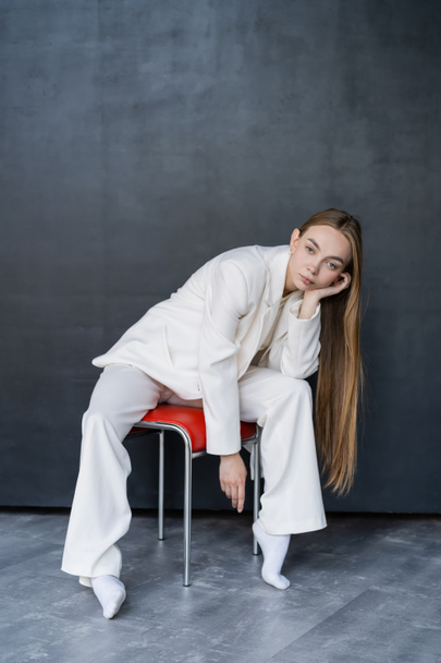 full length of stylish woman in white suit and socks sitting on chair on black background - Foto, Imagen