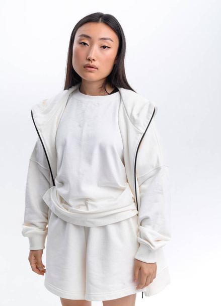 Una bella ragazza asiatica in una tuta bianca casual posa contro un muro bianco in uno studio fotografico - Foto, immagini