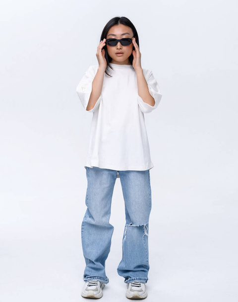 A beautiful Asian girl in a white t-shirt and blue jeans poses against a white wall in a photo studio. Fashion shooting - Foto, Imagem