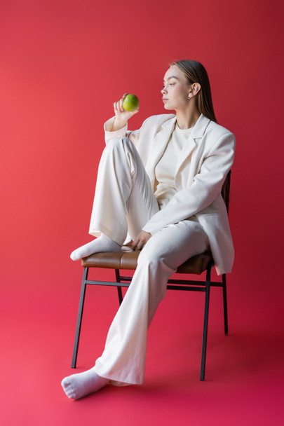 full length of woman in white suit and socks sitting on chair with apple on pink background - Photo, Image