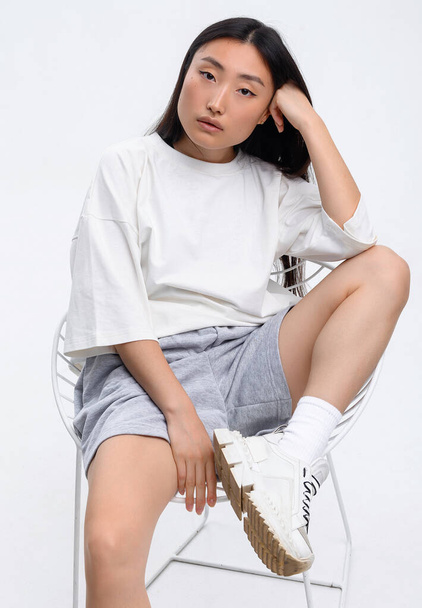 A beautiful Asian girl in a white t-shirt and shorts is posing against a white wall in a photo studio - Valokuva, kuva