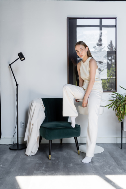 full length of woman in white trousers posing near black armchair and modern floor lamp - Foto, afbeelding
