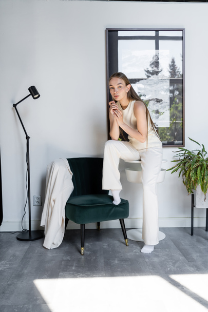 full length of woman in white clothes looking at camera near black armchair in modern room - Foto, afbeelding