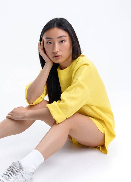 Прекрасна азіатська дівчина в жовтому лляному костюмі, що висить проти білої стіни у фотостудії. - Фото, зображення
