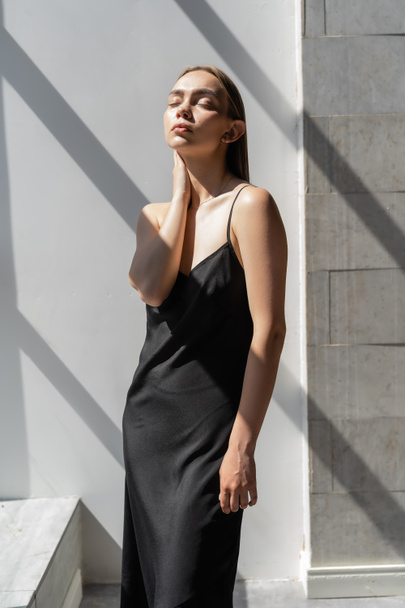 svůdná žena v černém popruh šaty stojící se zavřenýma očima a dotýkající se krku na bílém pozadí se stíny - Fotografie, Obrázek