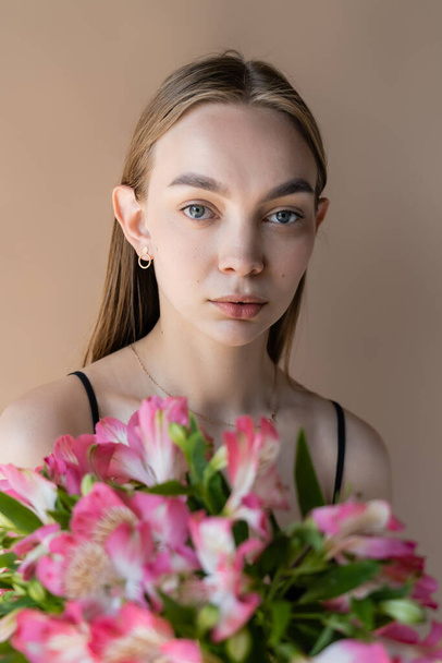 young woman with perfect skin looking at camera near alstroemeria flowers isolated on beige - Foto, immagini