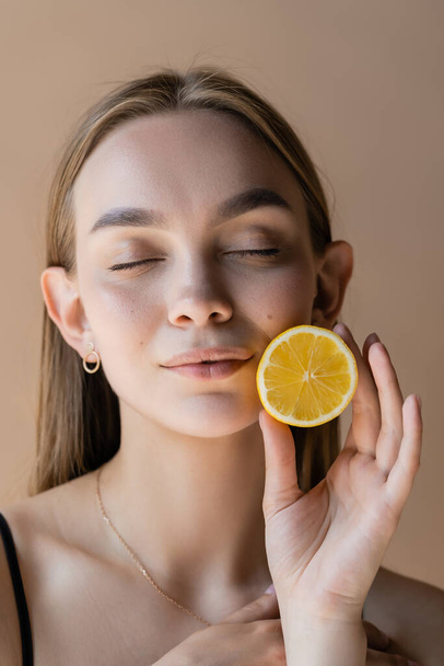 portrait of pleased woman with closed eyes holding half of juicy lemon isolated on beige - Fotó, kép
