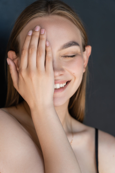 portrait of smiling woman with natural makeup obscuring face with hand isolated on black - Foto, imagen