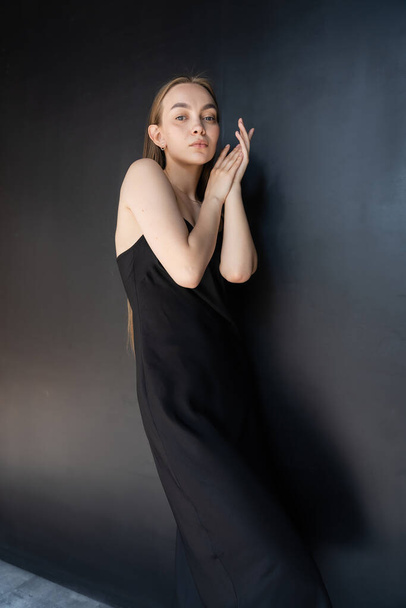 slim and seductive woman in black strap dress looking at camera near dark wall  - Zdjęcie, obraz