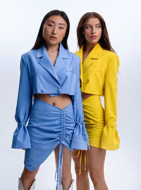 Two beautiful Caucasian and Asian girls lie on the white floor and pose in blue and yellow suits in a photo studio - Fotó, kép