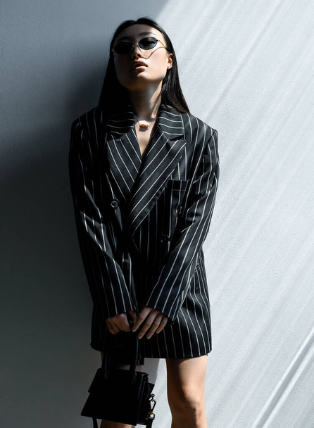 Beautiful Asian girl in black striped jacket wearing sunglasses posing against gray wall in photo studio. Fashion shooting - Fotoğraf, Görsel