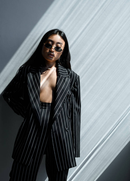 Beautiful Asian girl in black striped jacket wearing sunglasses posing against gray wall in photo studio. Fashion shooting - Photo, image