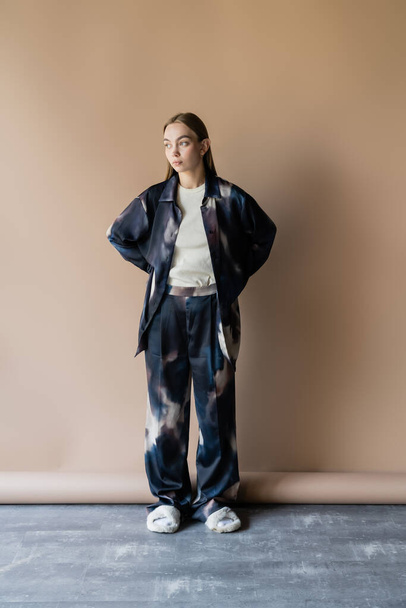 full length of woman in oversize clothes and soft slippers posing with hands behind back on beige background - 写真・画像