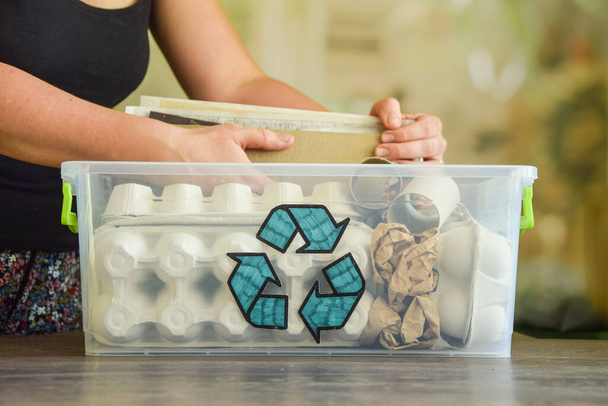 Female hand sorting waste paper. Transparent plastic container with recycle sign full of wastepaper on wooden table on blurred background. Recycling old paper products reused. Environment conservation - Φωτογραφία, εικόνα