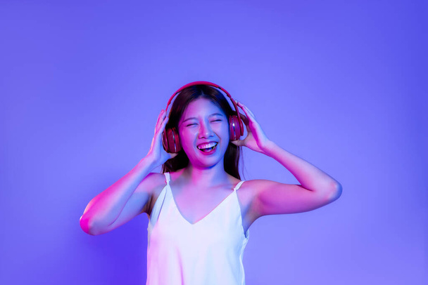 Young asian woman in white tank top wearing red headphone listen to music smiling on the purple neon light background.	 - Φωτογραφία, εικόνα