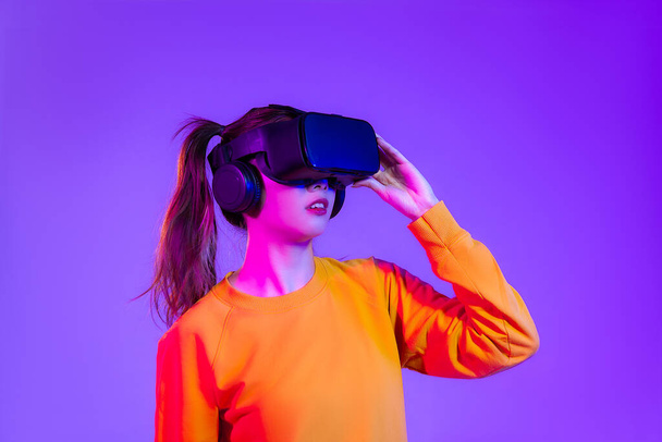 Metaverse concept. Young asian woman in orange sweatshirt wearing vr headset posing watching shopping and playing on the purple screen background.	 - Φωτογραφία, εικόνα