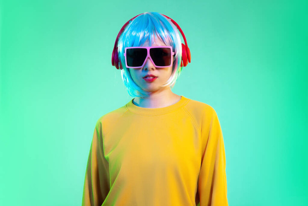 Young asian girl in yellow sweatshirt blue color shot hair style wearing red headphone and pink sunglasses listen to music posing standing on the green screen background. - 写真・画像