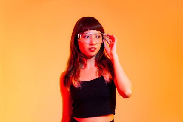 Young asian girl in black tank top wearing vr clear glasses posing on yellow screen background. - Photo, image