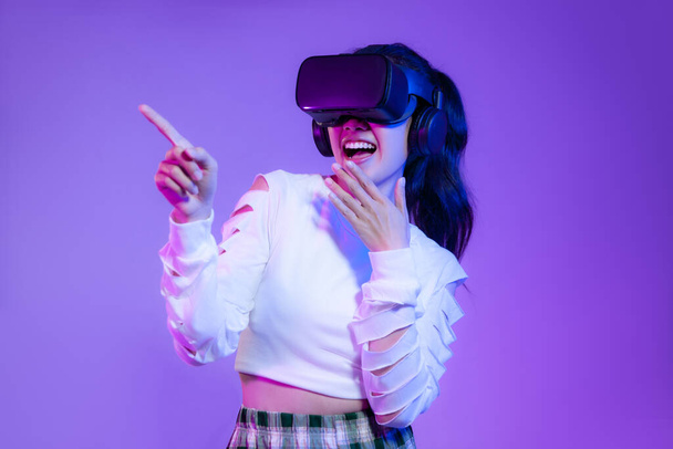 Young asian girl in wearing vr headset watching playing and finger pointing on the purple screen background.	 - Foto, imagen