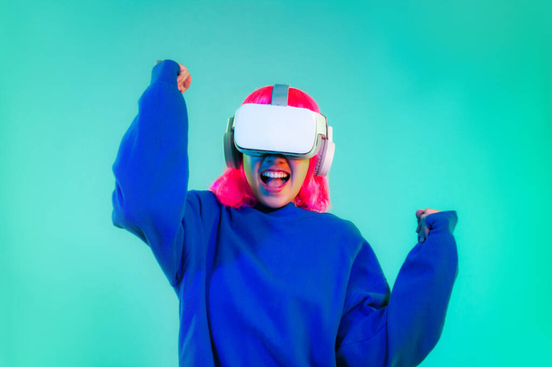 Young asian girl pink color hairstyle in blue sweatshirt wearing white vr headset watching playing cheering and touching on the green screen background. Cyborg and Metaverse concept.	 - Fotó, kép
