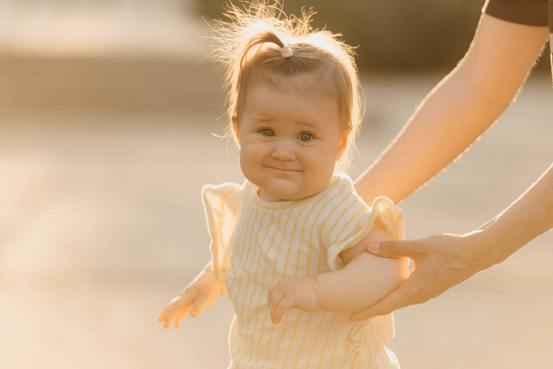 A close portrait of a female toddler at the hands of a mom in the sun lights. Mother is teaching her pretty young daughter to do first steps in the park at sunset. - Photo, Image