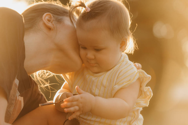 A female toddler in yellow clothes is playing with sunglasses near her mom in the sun rays. Mother is kissing her young daughter in the park at sunset. - Foto, Imagem