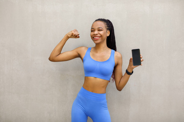 Black woman on gray background looking at camera while flexing biceps and holding phone. High quality photo - Zdjęcie, obraz
