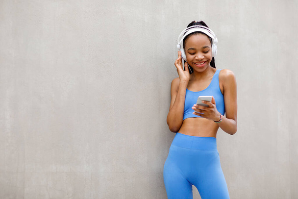Uśmiechnięta, muskularna, afrykańska młoda kobieta ze słuchawkami patrząca na smartfona na szarym tle, wolna przestrzeń. Wysokiej jakości zdjęcie - Zdjęcie, obraz
