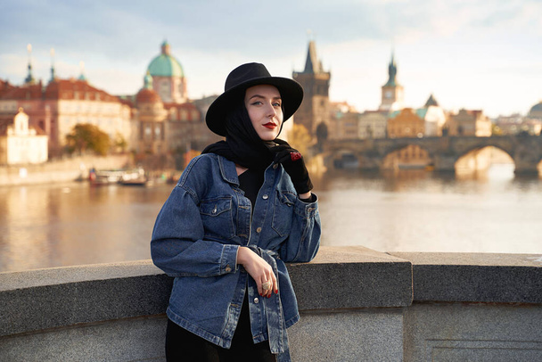 Stylish young beautiful woman earing black hat in Prague with Charles Bridge on background. Elegant retro lady fine art portrait. - Foto, imagen