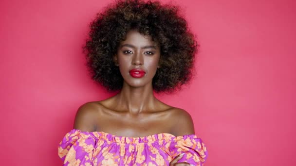 Mooie Afro-Amerikaanse vrouw met krullend haar en kale schouders kijken naar de camera en glimlachen op roze achtergrond - Video