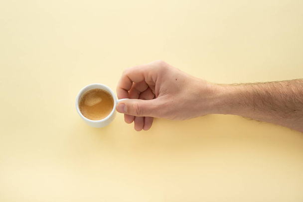Male hand holding a cup of espresso coffee on a yellow background. Morning coffee.  - 写真・画像