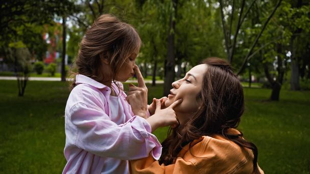 боковой вид счастливой матери, трогающей нос симпатичной дочери в зеленом парке - Фото, изображение