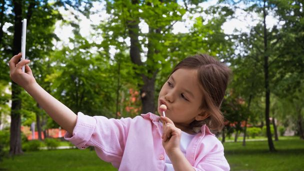bambino che tiene lecca-lecca sul bastone mentre prende selfie sullo smartphone nel parco  - Foto, immagini
