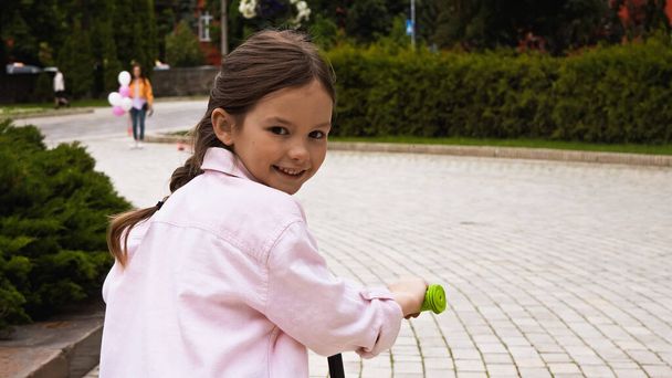 happy kid in pink denim jacket riding kick scooter in park  - Foto, Imagen